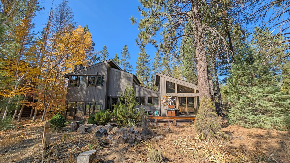 A modern architectural exterior featuring large windows and a wooden deck set among the trees. The image suggests a seamless flow between indoor and outdoor spaces, with room for both quiet contemplation and small gatherings. The deck overlooks lush greenery—perfect for watching golf action or simply enjoying the view.