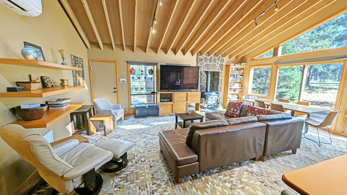 The image showcases a bright, inviting living room with expansive windows framing serene woodland views, a striking stone fireplace, exposed wooden beams, and plush, comfortable seating arranged for easy conversation. Decorative shelves add a personal, homey touch, while natural light bathes the space in warmth, creating an atmosphere perfect for both relaxation and gathering.