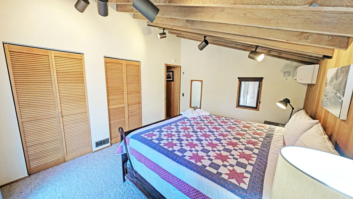 A cozy bedroom featuring a queen bed with a quilted spread, rustic wooden beams overhead, closet doors for ample storage, and thoughtfully placed artwork on the wall. Gentle natural light adds warmth to the restful setting.