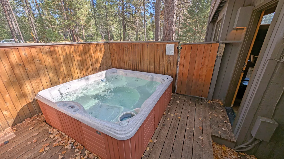 A wide view of the wooden deck with a bubbling hot tub as the centerpiece, surrounded by mature trees and with the entrance door visible. The peaceful, natural setting is ideal for relaxing, socializing, or enjoying a quiet retreat after a day on the golf course.