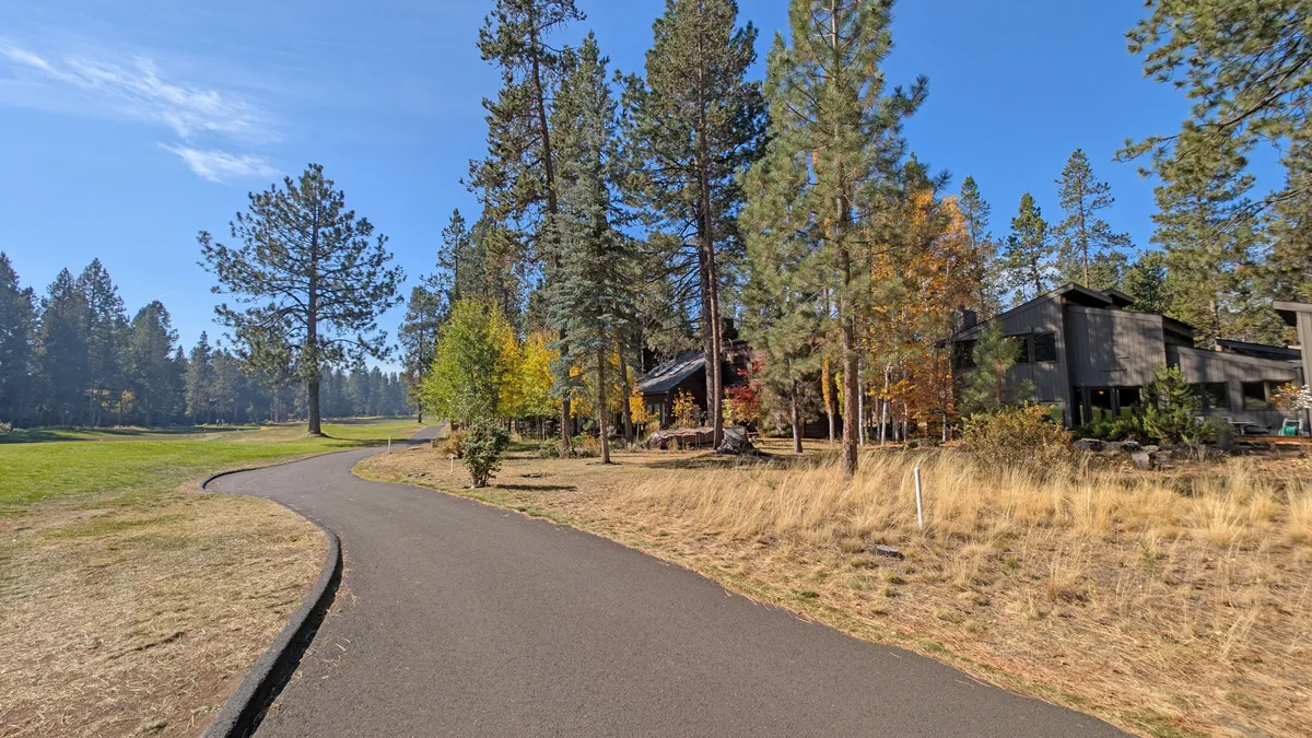 A scenic exterior view featuring a winding path surrounded by tall pine trees and vibrant, colorful foliage, with modern architecture visible in the background. The image evokes a sense of outdoor adventure and connection to nature, perfectly matching the Sunriver experience.