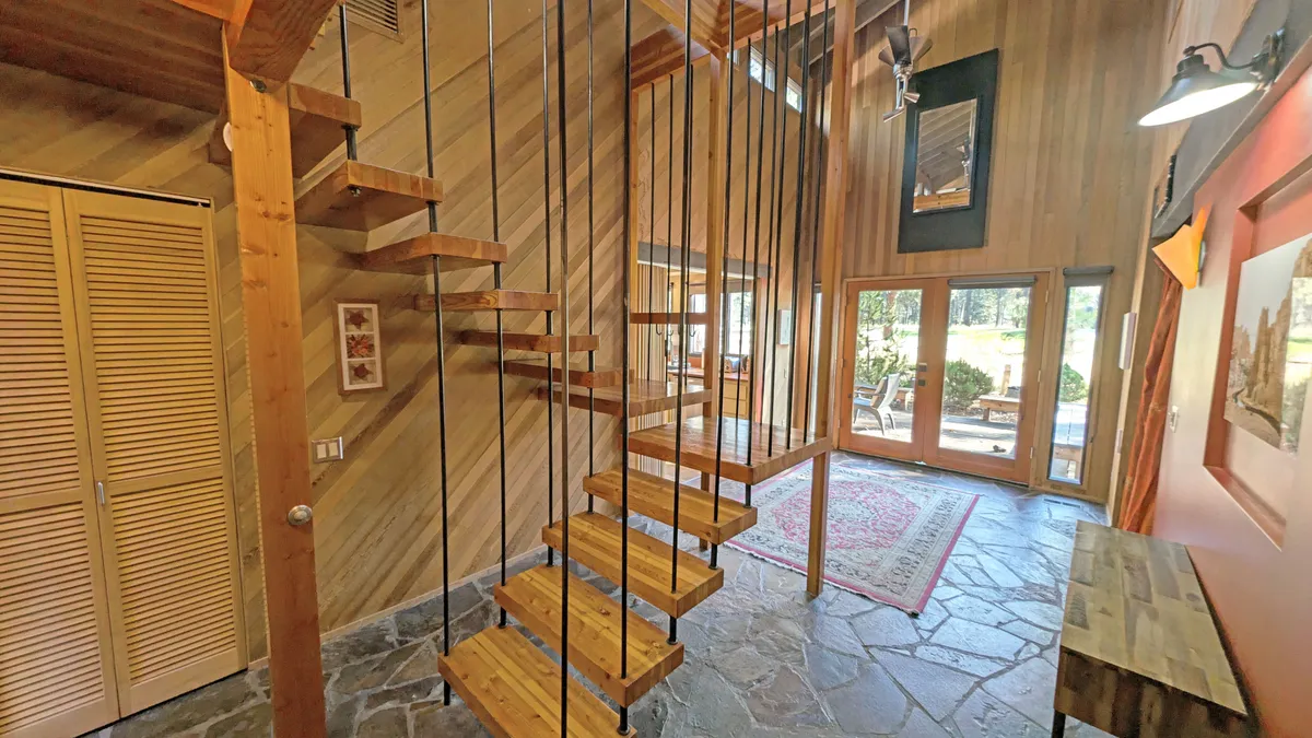 A floating staircase with sleek lines, slate flooring, large windows, wooden walls, and decorative artwork sets a modern, open, and airy mood. The space is designed for welcoming guests and family gatherings while maximizing natural light and showcasing contemporary architectural elements.