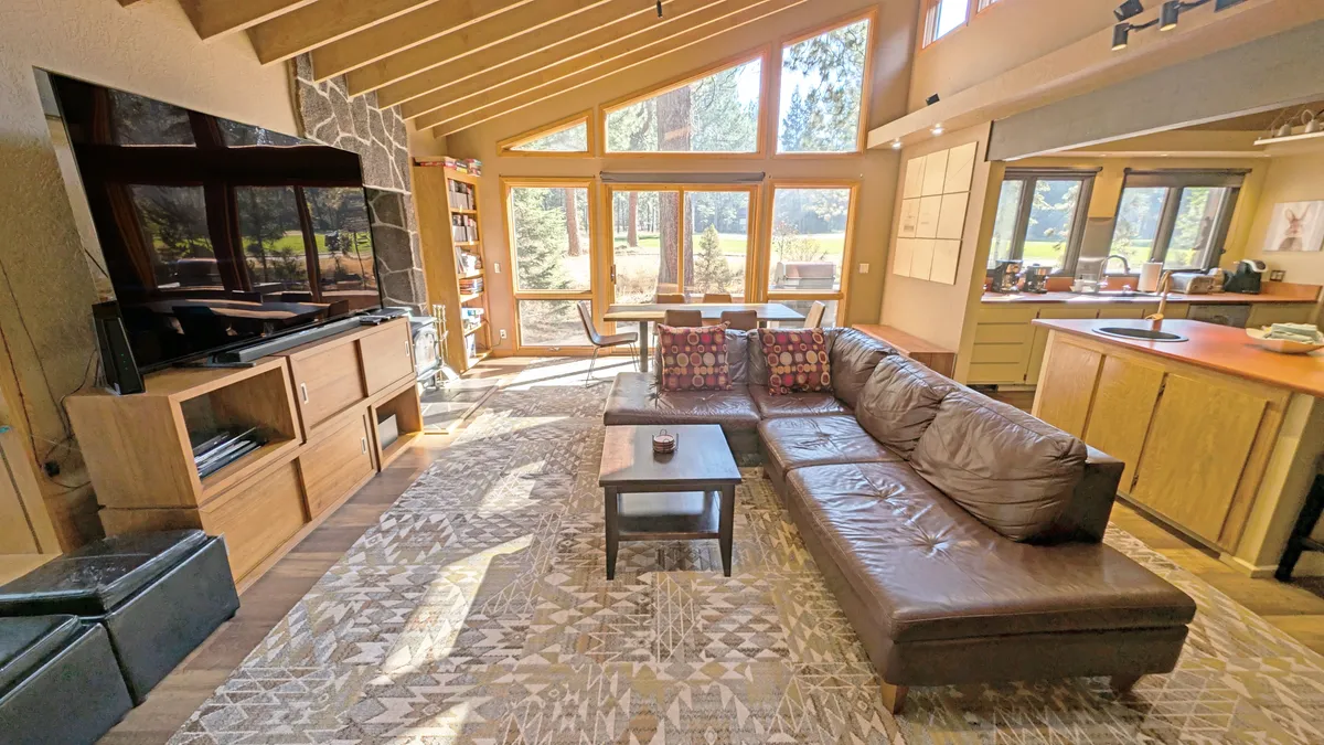 A welcoming open-plan living and dining area featuring a comfortable leather sofa, a contemporary TV unit, decorative rug, and a wooden dining table, all set against the backdrop of expansive windows offering views of nature and letting in brilliant sunlight.