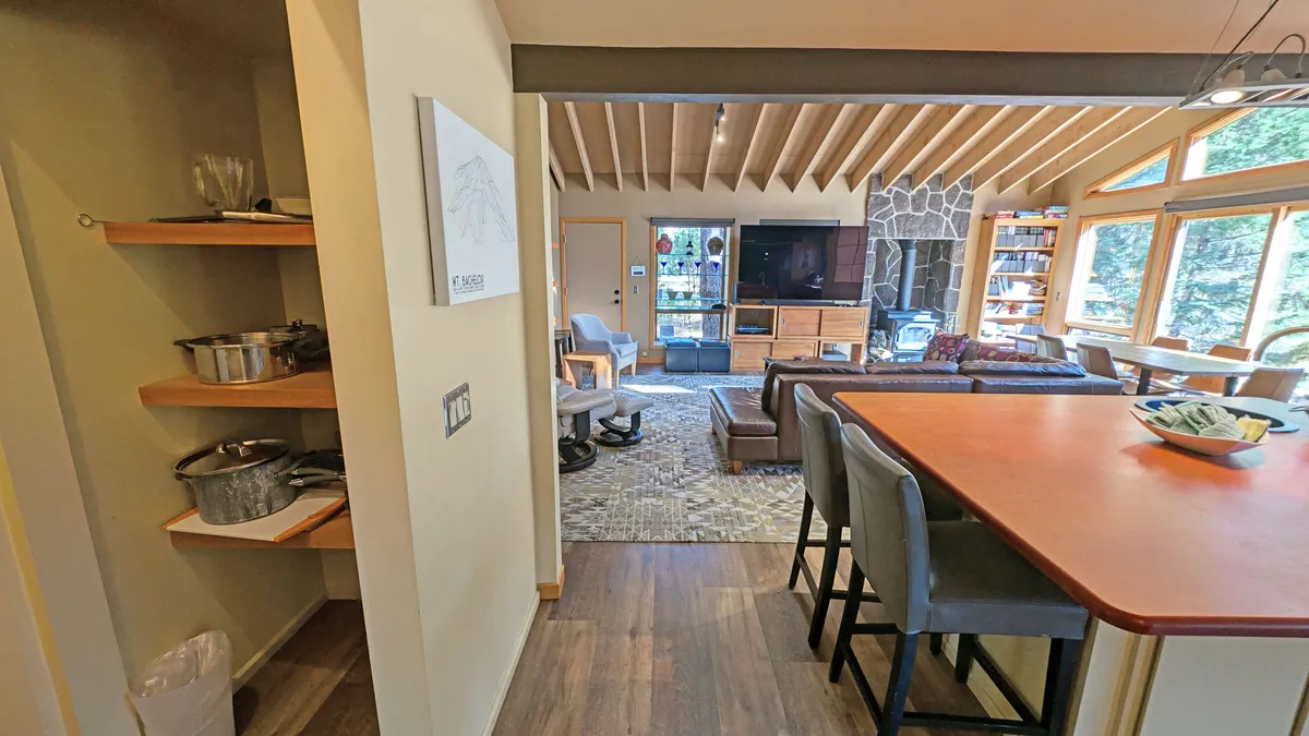 A spacious, inviting great room showcasing an open layout with a stone fireplace, large windows, a kitchen island, and decorative shelves. The area is designed for socializing, relaxing, and entertaining—perfect for group gatherings.