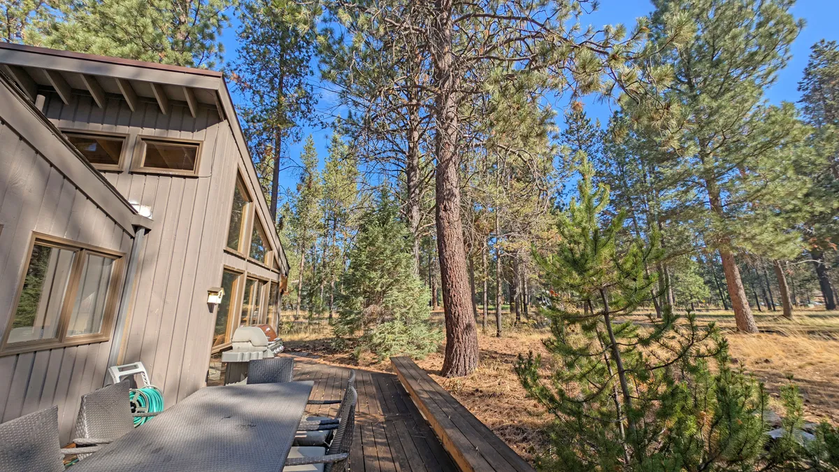 A peaceful wooden deck nestled among the trees, featuring a table with chairs for outdoor dining or gathering. The surrounding greenery creates an inviting atmosphere for family BBQs, morning coffee, and evenings spent under the stars.