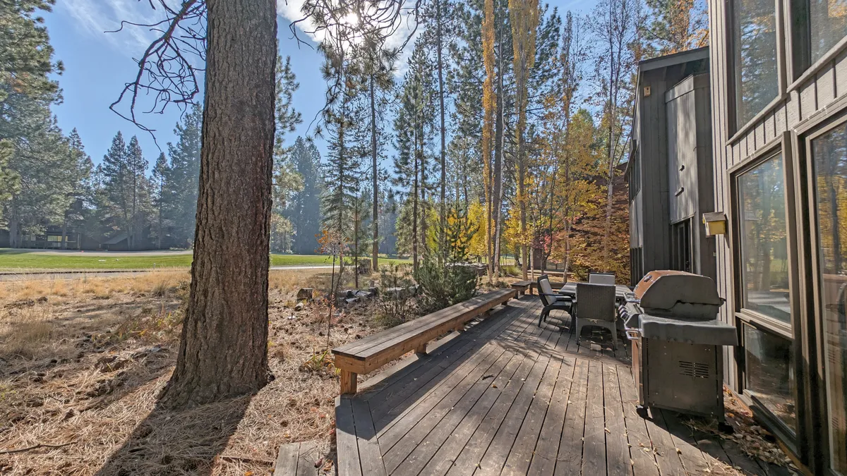 This image shows a wooden deck furnished with a grill, surrounded by tall trees and natural greenery. There's a pathway leading away from the deck, inviting outdoor exploration, and the setting feels peaceful and connected to nature.