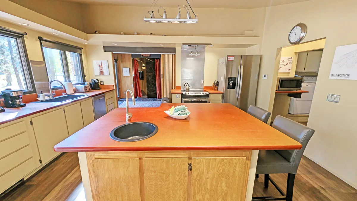 A bright, modern kitchen with a spacious central island, large windows that flood the area with natural light, stainless steel appliances, and warm wooden cabinetry. The open design invites easy movement between the kitchen and adjacent gathering spaces, all set within an inviting, functional atmosphere.