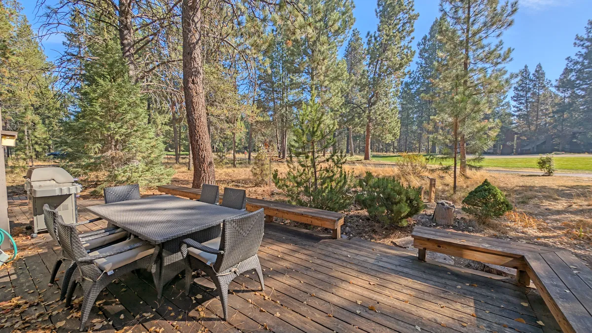 A spacious outdoor dining area with a wooden table and chairs sits on a deck nestled among tall trees. An outdoor grill is ready for barbecues, making this an inviting setting for alfresco meals, gatherings, and tranquil moments in nature.