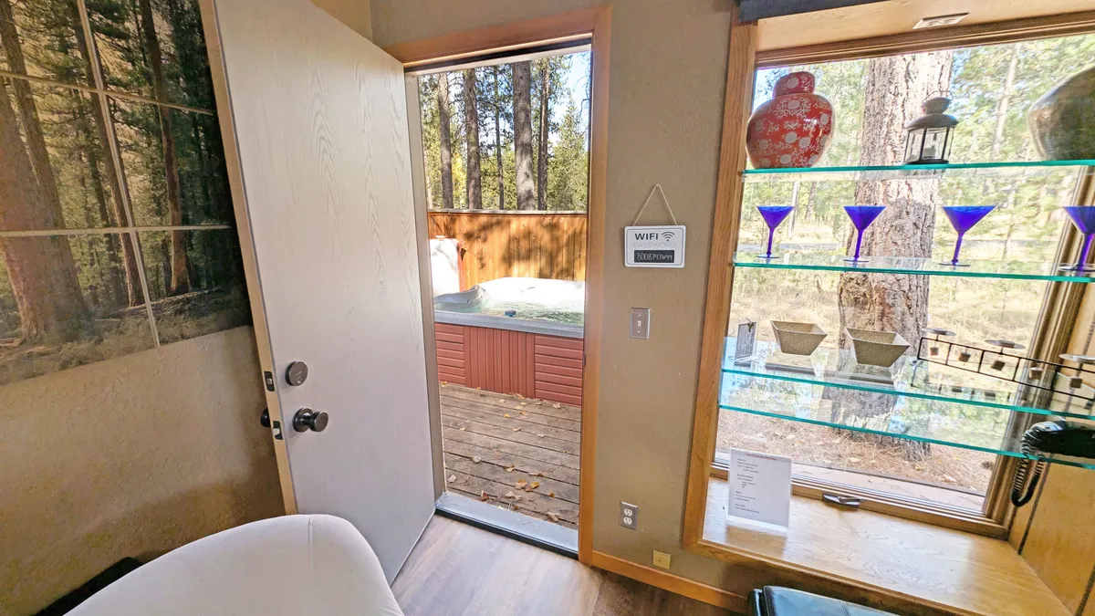 This image showcases a tranquil outdoor deck area with a private hot tub, surrounded by lush forest views. A door opens directly onto the deck, seamlessly connecting the indoor living space to the outdoor sanctuary. Decorative glassware adds a pop of color to a glass shelf inside, hinting at the ease of indoor-outdoor living.