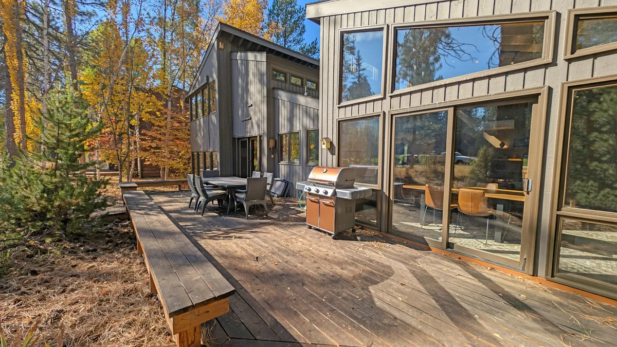 An inviting outdoor dining area set on a spacious deck, surrounded by tall trees and featuring a grill. Large windows connect the interior to the vibrant natural setting, suggesting easy access to outdoor adventure and relaxation.