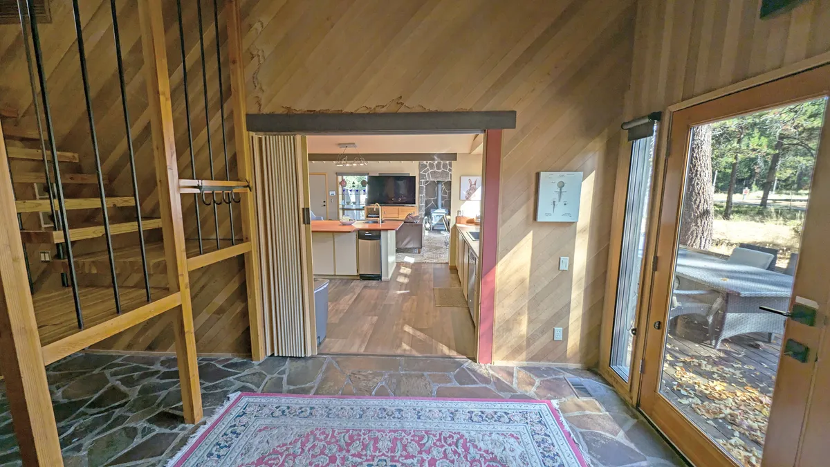 This image highlights an open kitchen adjacent to a wooden staircase, with large windows offering views of the surrounding landscape and a cozy stone wall. The kitchen and living areas are unified, emphasizing the home's updated, communal, and nature-connected design.