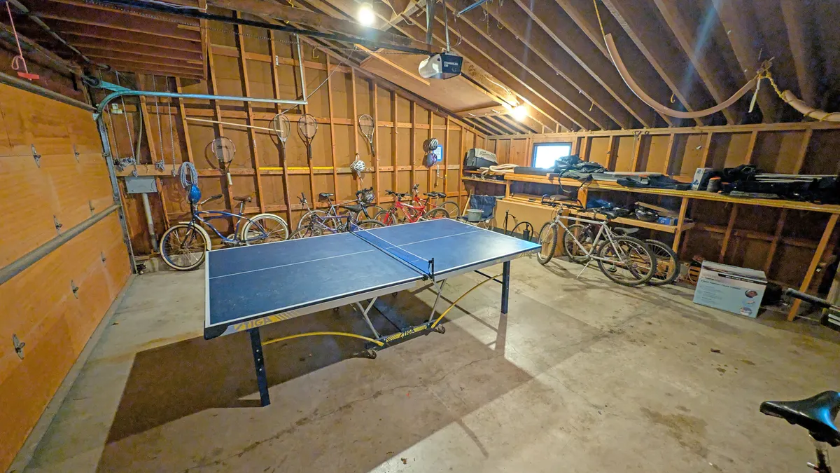 The image depicts a garage space that includes a ping pong table and several bicycles lined up along the wall, with storage shelves visible in the background. The area feels organized yet playful, emphasizing both recreation and practical storage for active gear.